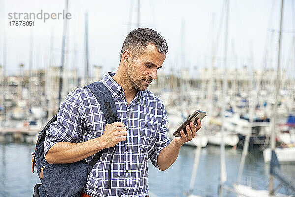 Reifer Mann mit Rucksack benutzt Smartphone im Hafen
