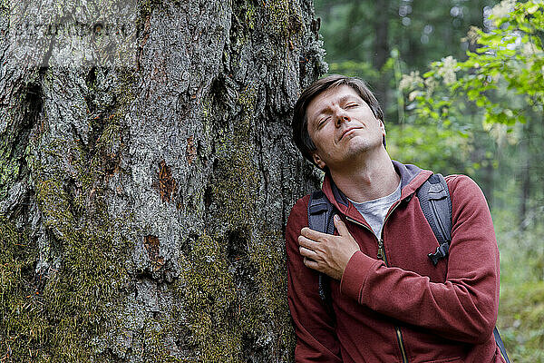 Mann mit geschlossenen Augen lehnt an Baum im Wald