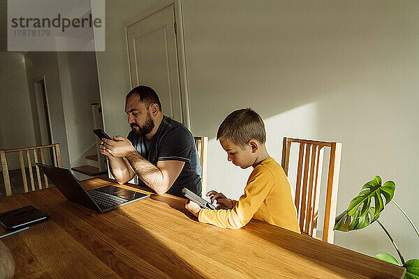 Mann benutzt Smartphone und sitzt zu Hause neben seinem Sohn mit Tablet-PC