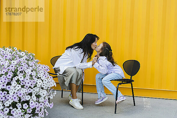 Zärtliche Mutter küsst Tochter vor gelber Wand auf den Mund