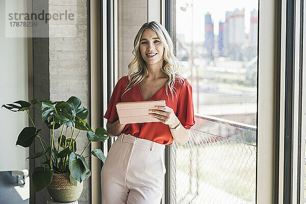 Lächelnde blonde Geschäftsfrau mit Tablet-PC lehnt am Fenster im Büro