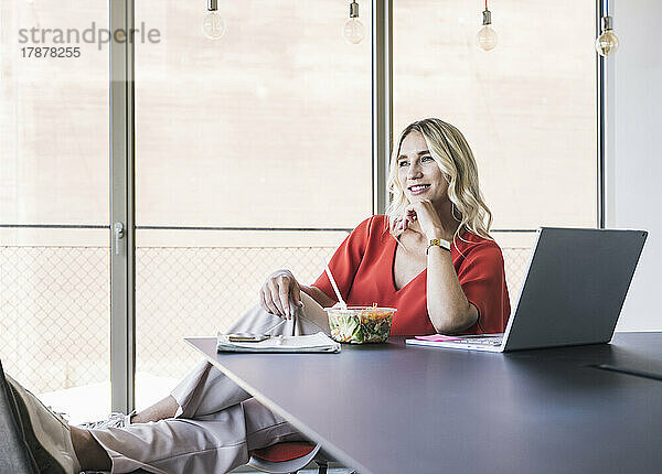 Nachdenkliche Geschäftsfrau mit der Hand am Kinn sitzt am Schreibtisch im Büro