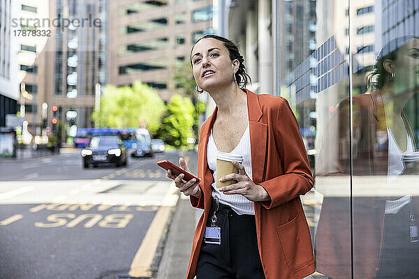 Geschäftsfrau hält Einwegbecher und Smartphone an Glaswand