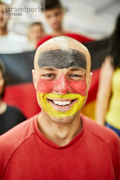 Junger lächelnder Mann mit bemaltem Gesicht mit deutscher Flagge im Stadion