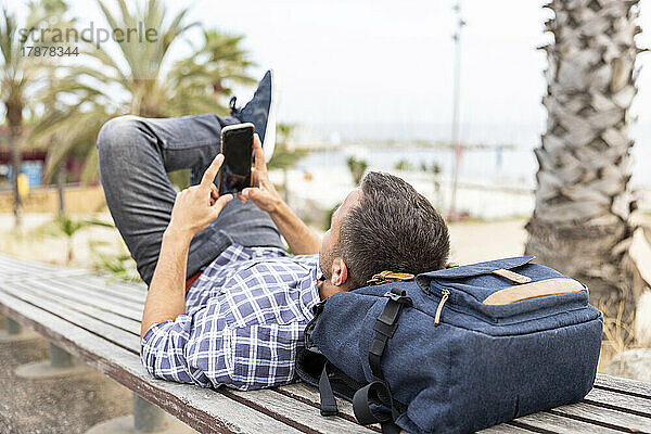 Reifer Mann benutzt Smartphone und entspannt sich auf einer Bank am Strand