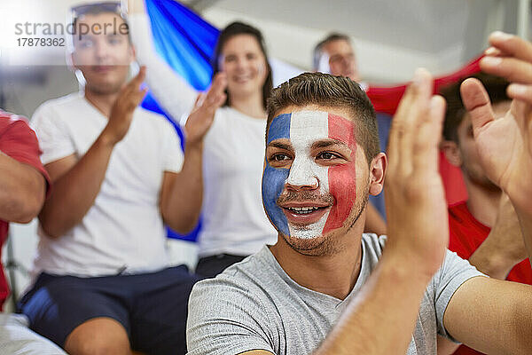 Nachdenklicher Mann mit aufgemalter französischer Flagge im Gesicht klatscht mit Fans im Stadion