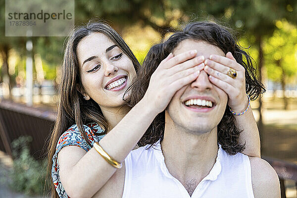 Glückliche junge Frau bedeckt ihrem Freund im Park die Augen