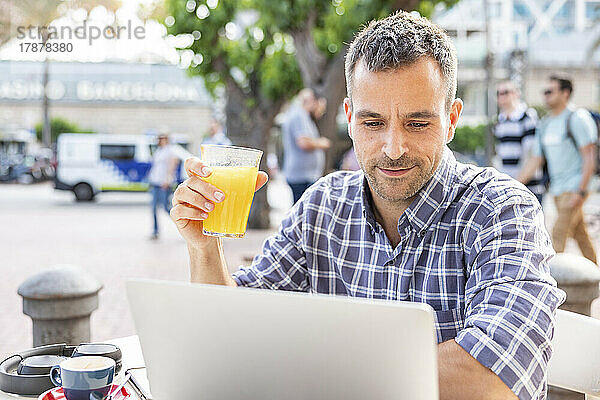 Reifer Mann arbeitet am Laptop und trinkt Orangensaft im Straßencafé