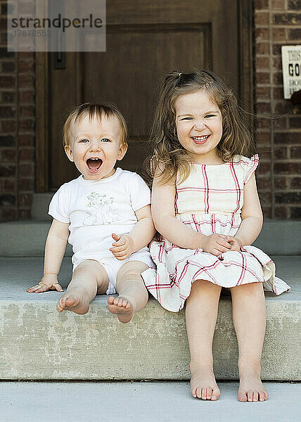 Porträt eines lächelnden Bruders (12–17 Monate) und einer Schwester (2–3)  die auf der Veranda sitzen