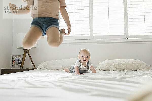 Mädchen (2-3) springt auf Bett  im Hintergrund ihr kleiner Bruder (6-11 Monate).