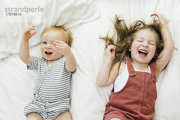 Draufsicht auf Bruder (12–17 Monate) und Schwester (2–3)  die im Bett lachen