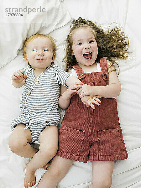 Draufsicht auf Bruder (12–17 Monate) und Schwester (2–3)  die im Bett lachen