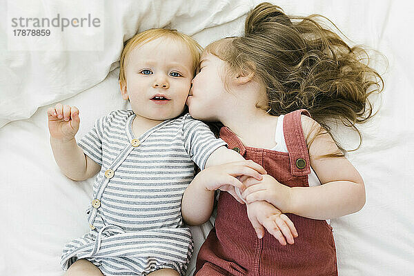 Draufsicht auf ein Mädchen (2–3)  das seinen Bruder (12–17 Monate) auf dem Bett küsst