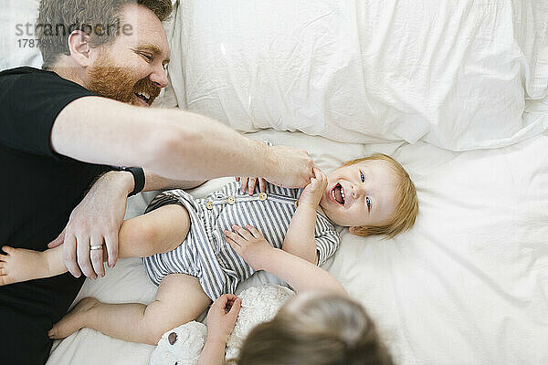 Vater spielt mit Sohn (12–17 Monate) und Tochter (2–3) im Bett