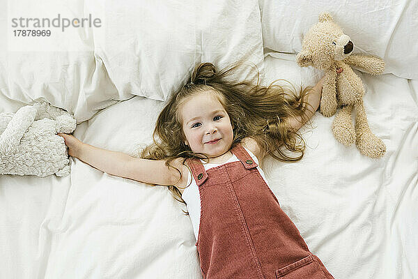 Blick von oben auf ein lächelndes Mädchen (2-3)  das mit Stoffspielzeugen auf dem Bett liegt