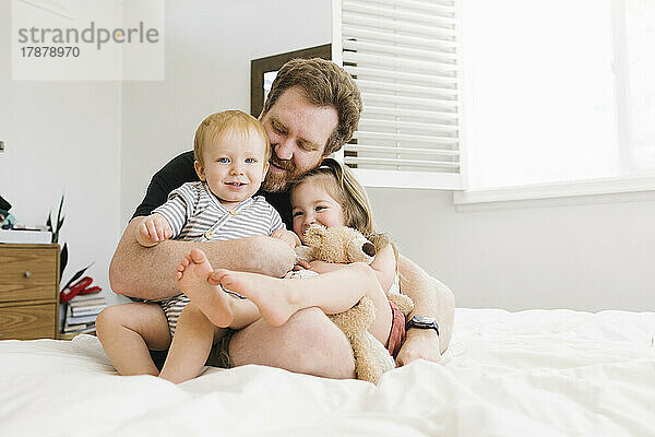 Vater umarmt Sohn (12–17 Monate) und Tochter (2–3) im Bett