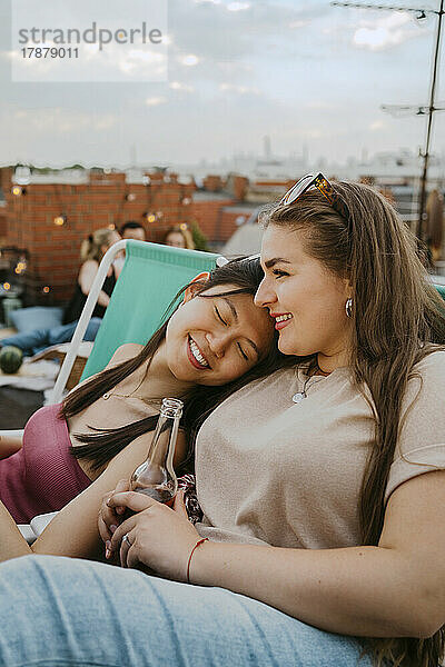 Lächelnde junge Frau mit einer Bierflasche in der Hand  die mit geschlossenen Augen auf einem Dach bei ihren Freundinnen sitzt