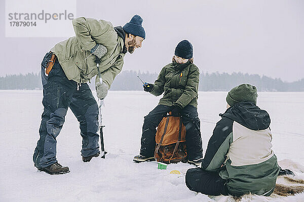 Fröhlicher Mann  der einen Eisbohrer benutzt und sich mit einem Rucksack auf einem zugefrorenen See unterhält