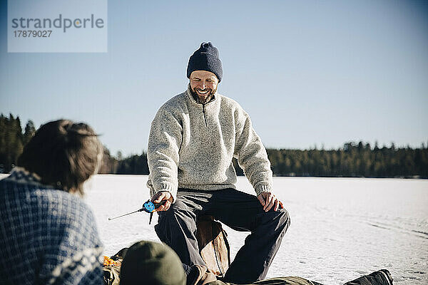 Glücklicher reifer Mann mit Angelrute  der mit Freunden an einem sonnigen Tag im Winter sitzt
