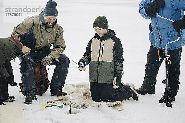 Glücklicher Junge kniet neben Männern und einem Freund beim Angeln auf einem zugefrorenen See im Winter
