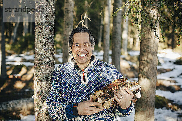 Porträt eines glücklichen reifen Mannes mit Brennholz an einem Baum im Wald