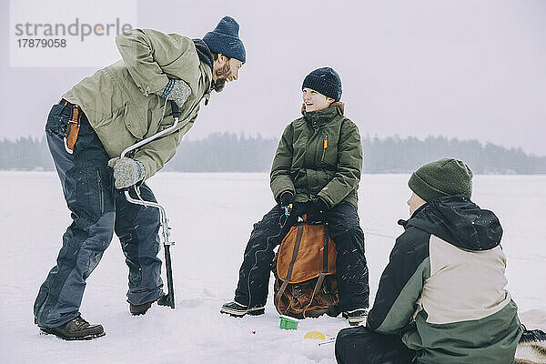 Glücklicher reifer Mann  der einen Eisbohrer benutzt  während er sich mit seinem Sohn unterhält  der auf einem Rucksack auf einem zugefrorenen See sitzt