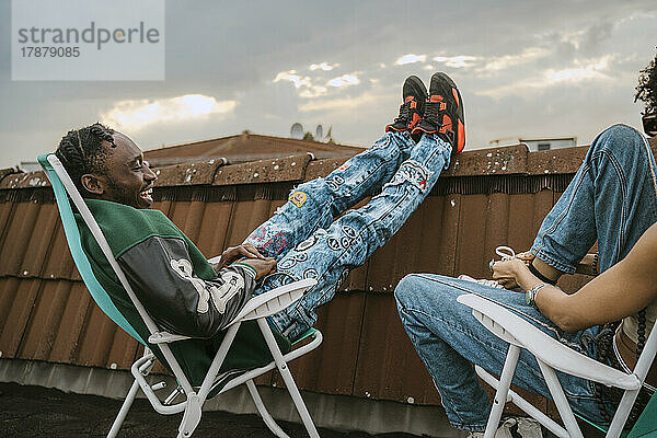Seitenansicht eines fröhlichen jungen Mannes  der sich mit einer Freundin unterhält  während er auf einem Stuhl auf einer Dachterrasse sitzt