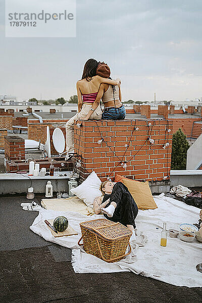 Frau auf Decke liegend mit Freundinnen  die auf dem Dach sitzen und sich umarmen