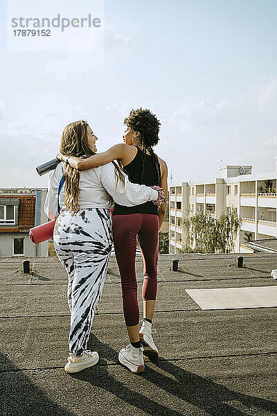 Rückansicht von weiblichen Freunden  die auf einem Dach in der Sonne spazieren gehen und sich umarmen