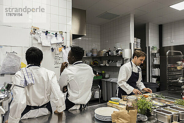 Multirassische Köche diskutieren am Whiteboard bei der Zubereitung von Speisen in einer Großküche