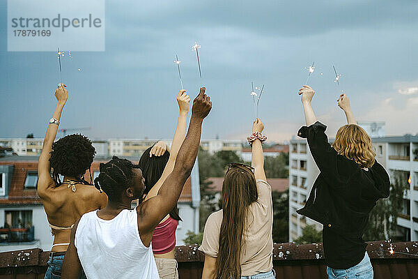 Rückansicht von multirassischen Freunden mit angezündeten Wunderkerzen und erhobenen Händen auf dem Dach