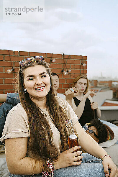 Porträt einer lächelnden jungen Frau  die eine Bierflasche in der Hand hält und vor Freunden auf einem Dach sitzt