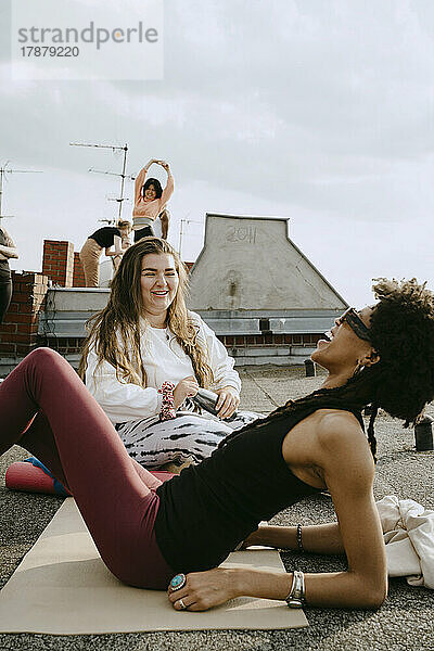Lächelnde junge Frau im Gespräch mit einer fröhlichen Freundin auf einer Übungsmatte liegend