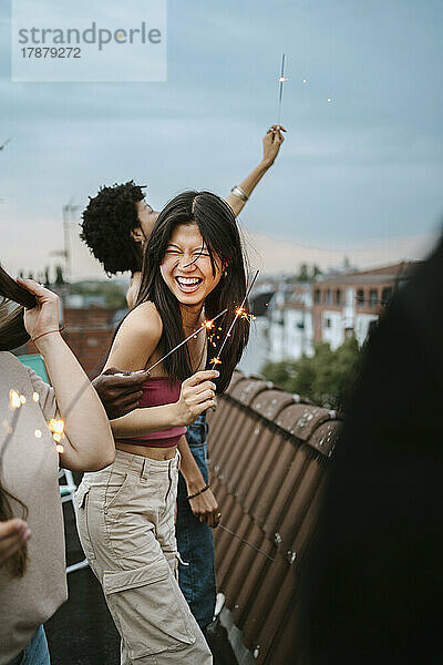 Fröhliche junge Frau  die eine Wunderkerze in der Hand hält und sich mit Freunden auf einem Dach vergnügt