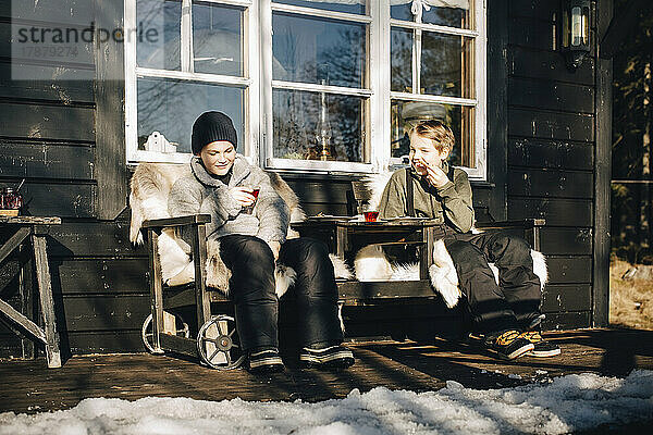 Lächelnde Freunde in voller Länge auf Stühlen sitzend auf einer Veranda im Winter