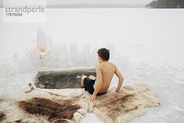 Rückansicht eines Jungen ohne Hemd  der mit den Beinen im kalten Wasser eines zugefrorenen Sees sitzt