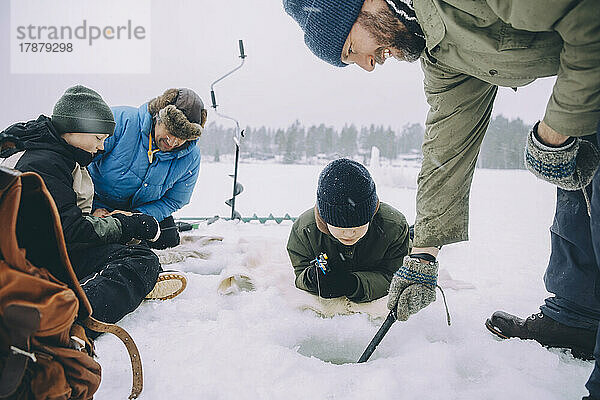 Junge sieht Vater beim Eisfischen zu  während Freunde im Urlaub im Schnee sitzen