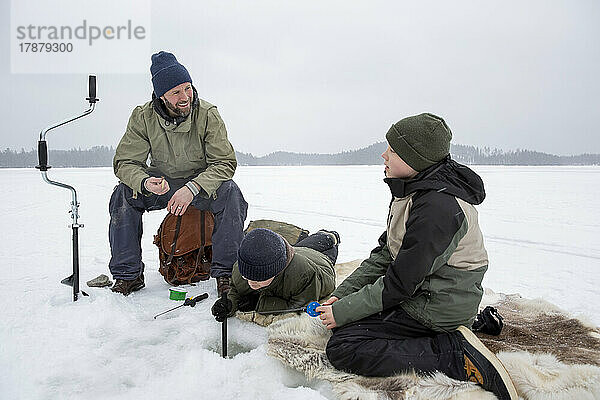 Lächelnder Mann im Gespräch mit einem Jungen  während sein Sohn auf einem zugefrorenen See Eisfischen geht