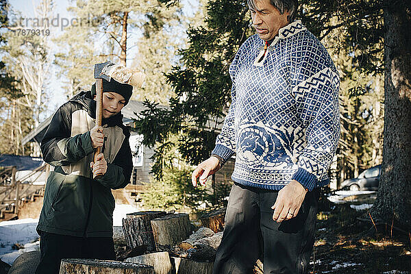 Älterer Mann sieht seinen Sohn an  der im Winter im Wald einen Baumstamm mit einer Axt schneidet