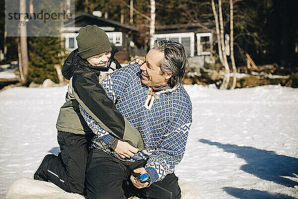 Glücklicher Junge in warmer Kleidung umarmt seinen Vater beim Eisfischen an einem sonnigen Tag