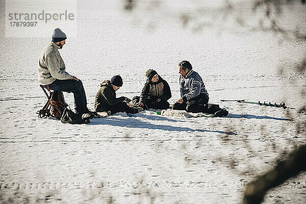 Ältere Männer mit Söhnen sitzen beim Eisfischen im Schnee bei sonnigem Wetter