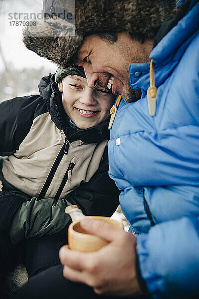 Glücklicher Vater  der eine Tasse Tee hält und seinen Sohn in warmer Kleidung umarmt