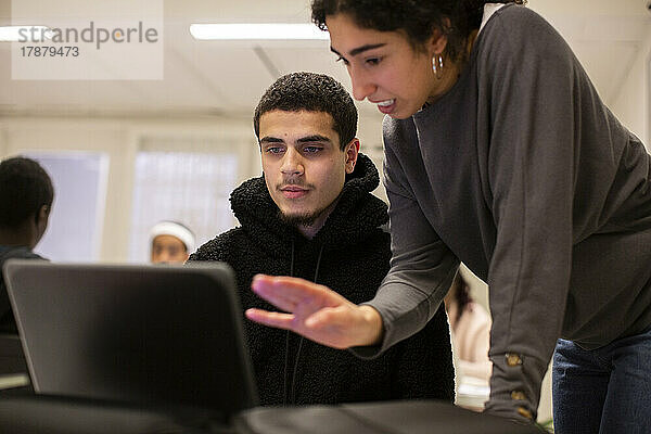 Schülerin schaut auf den Laptop  während der Lehrer im Klassenzimmer etwas erklärt