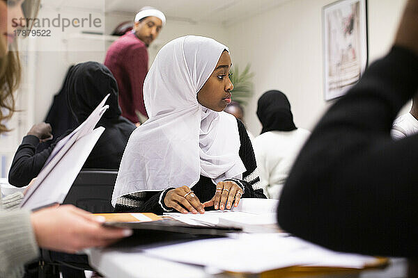 Mädchen im Hidschab sitzt mit Freunden am Schreibtisch im Klassenzimmer