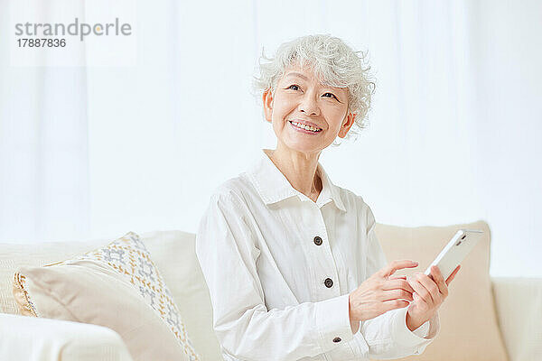 Japanische Seniorin mit Smartphone auf dem Sofa