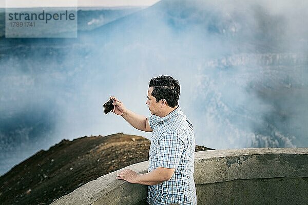 Tourist  der an einem Aussichtspunkt Fotos macht. Junger Tourist  der an einem vulkanischen Aussichtspunkt Fotos macht. Abenteuerlustiger Mann mit seinem Mobiltelefon  der an einem Aussichtspunkt Fotos macht. Volcán Masaya  Nicaragua  Mittelamerika