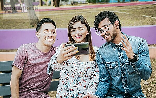Nahaufnahme von drei Freunden  die im Park sitzend ein Selfie machen  Drei lächelnde Freunde  die im Park sitzen und ein Selfie machen. Vorderansicht von drei glücklichen Freunden  die ein Selfie machen  während sie auf einer Bank sitzen