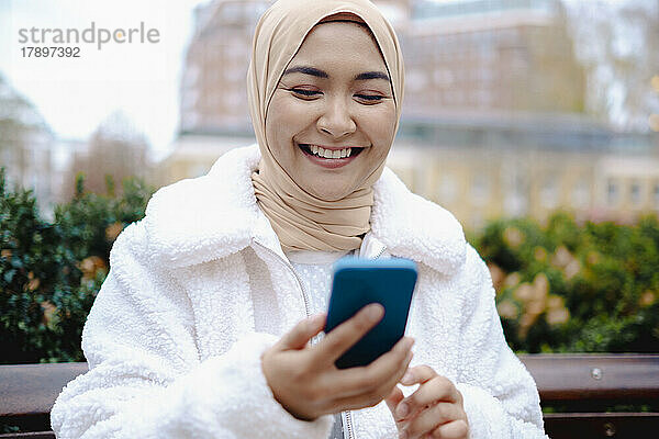 Glückliche junge Frau mit Hijab  die im Park ihr Mobiltelefon benutzt