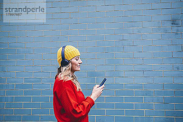Junge Frau mit kabellosen Kopfhörern und Smartphone vor der Wand