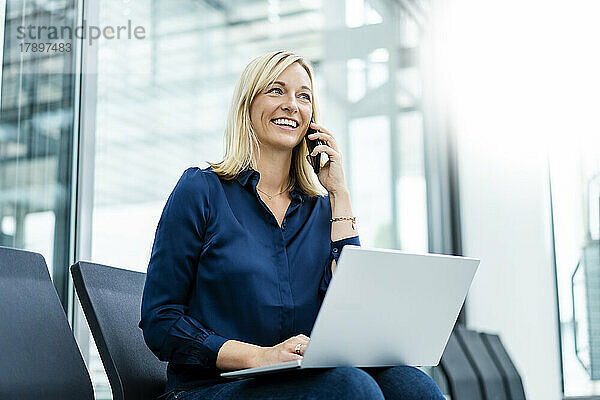 Lächelnde Geschäftsfrau mit Laptop  die in der Lobby telefoniert
